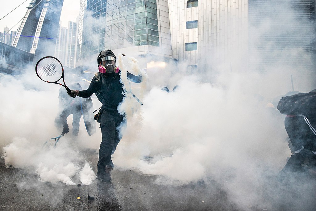 Image of masked protester using tennis racket to bat away tear gas