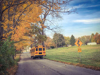 Picture of a school bus driving down a rural road with a 20 mile per hour sign and fall foliage